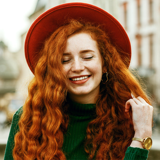 Mujer pelirroja con cabello largo y ondulado, sonriendo. Ideal para productos de cuidado capi - Luxent Professional