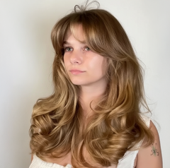 Mujer con cabello rubio miel, flequillo cortina y ondas voluminosas. Peinado moderno y saluda - Luxent Professional